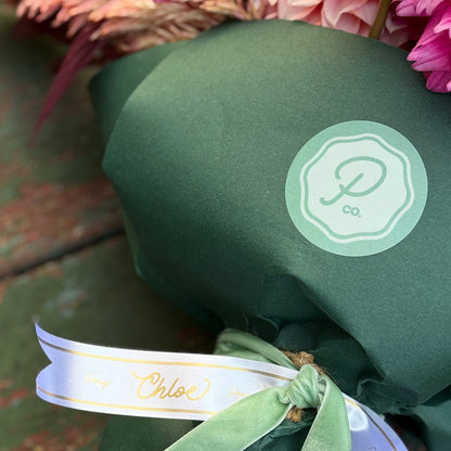 A close-up of a wrapped flower bouquet with pink and peach blossoms, green foliage, and a dark green paper wrap tied with a white ribbon. A circular mint-green logo sticker is visible on the wrap.
